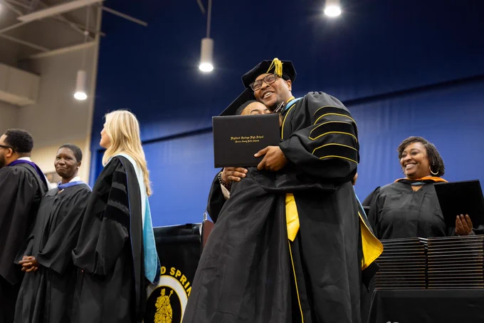 Several people in graduation gowns and caps on stage, with one person embracing another amidst crowd.