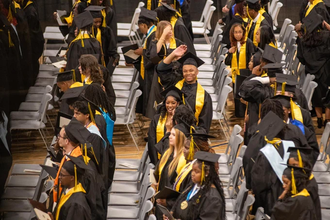 Graduates in black and gold gowns walking in procession at a commencement ceremony.