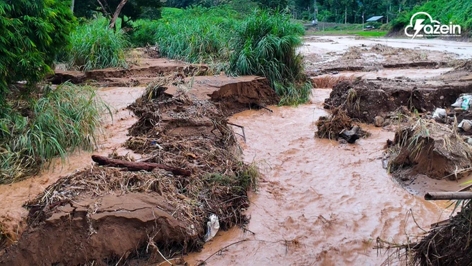 A landscape photograph showcasing severe soil erosion and flooding caused by heavy rainfall. A fast-moving, muddy brown river cuts through a landscape, carving deep into the dark soil and washing away parts of the embankment. Thick clumps of tall green grass and vegetation hang over the newly eroded, collapsed dirt banks, while scattered logs and plant debris are strewn across the center of the rushing water. The white "azein" logo is visible in the top right corner.