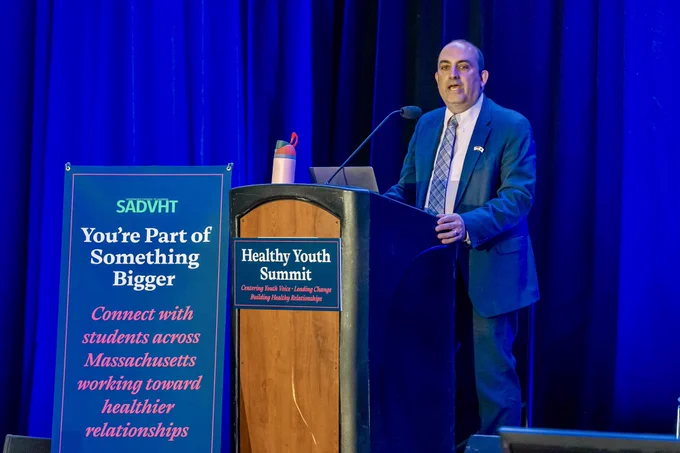 Education Secretary Steve Zrike speaks at a podium during the Healthy Youth Summit. A sign beside the stage reads, “You’re Part of Something Bigger — Connect with students across Massachusetts working toward healthier relationships.”