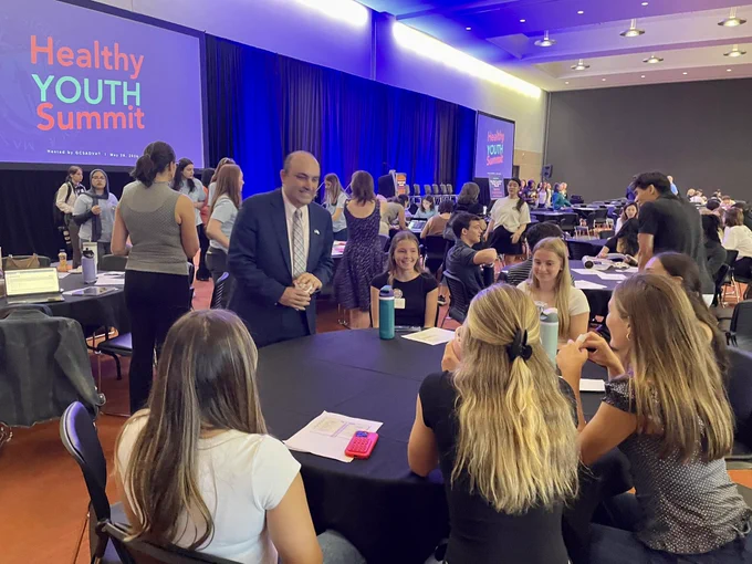 Education Secretary Steve Zrike stands in a suit and speaks to a group of students seated a table during the Healthy Youth Summit. Students and attendees sit at round tables listening as blue stage lighting fills the conference hall.