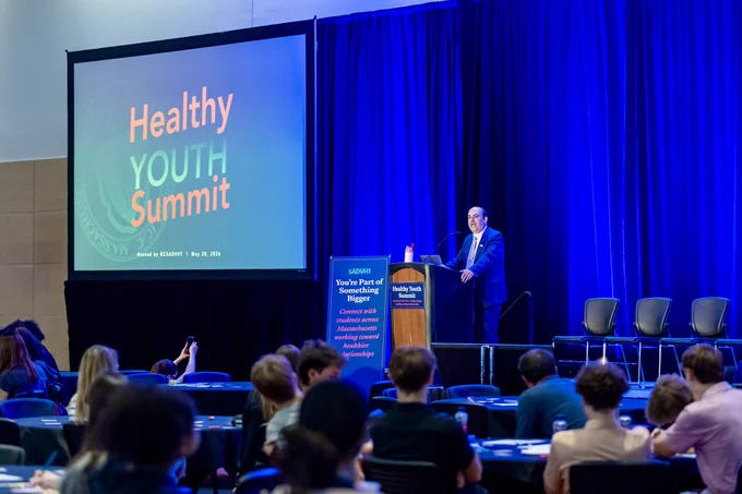 Education Secretary Steve Zrike speaks at the Healthy Youth Summit from a stage in front of a large screen displaying the event logo. Students and attendees sit at round tables listening as blue stage lighting fills the conference hall.