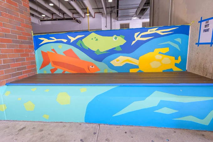 Vibrant mural with geometric fish in red, green, and yellow hues on a seating area. Background features bold blues and abstract aquatic shapes. Energetic and lively tone.