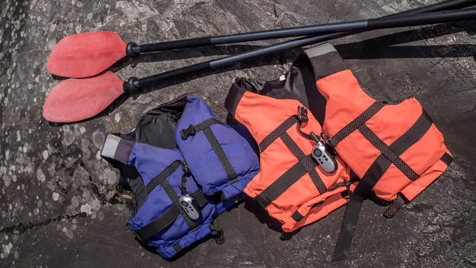Orange and blue lifejackets laid out on a rocky surface with two black and red paddles.