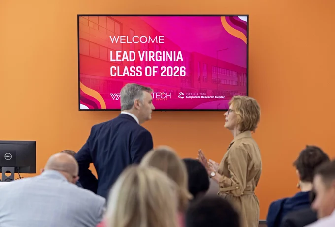 Two people chat in front of a display screen with "WELCOME LEAD VIRGINIA CLASS OF 2026"