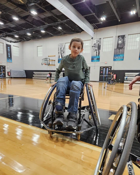 A young boy in a wheelchair on a basketball court, smiling. Other wheelchair users are in the background.