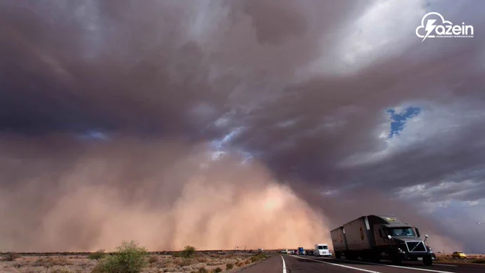 A dramatic photograph of a massive wall of dust, or haboob, rolling across a desert landscape toward a highway. In the foreground, a semi-truck with a trailer and a line of other vehicles are driving down a multi-lane road under an overcast sky filled with heavy, dark storm clouds. The giant cloud of tan dust towers over the flat desert terrain on the left, severely reducing visibility in the distance. The white "azein" logo is visible in the top right corner.
