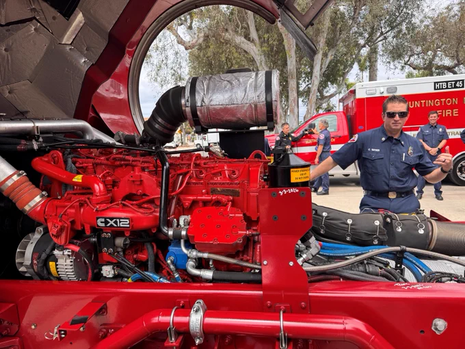 A Huntington Beach Fire Department firefighter stands beside an open fire engine compartment during the annual apparatus inspection. The engine's red Cummins powerplant and mechanical systems are fully exposed, allowing inspection of critical components. Additional HBFD vehicles and personnel can be seen in the background as crews conduct detailed evaluations to ensure apparatus reliability and readiness for emergency response.