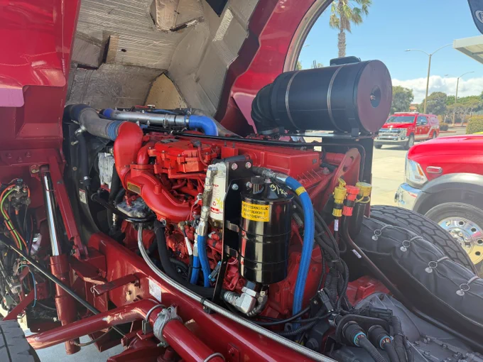 Close up view of a Huntington Beach Fire Department fire engine with the rear engine compartment opened during the annual apparatus inspection. The engine, hoses, filters, and mechanical systems are visible as personnel inspect and evaluate equipment condition and functionality. Department vehicles can be seen in the background, highlighting the ongoing effort to maintain a safe and reliable emergency response fleet.