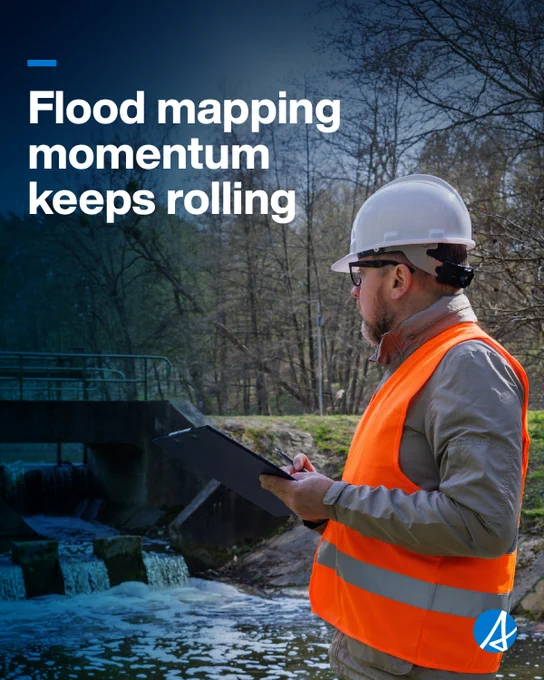 A man wearing a hard hat and orange safety vest is holding a clipboard and standing next to a river. Text reads Flood mapping momentum keeps rolling.
