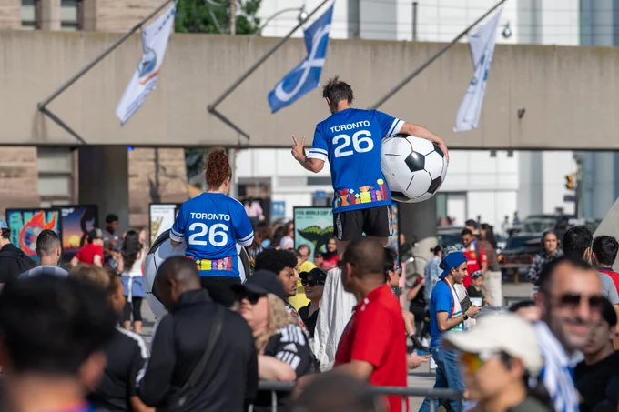 Two performers wearing blue Toronto 26 jerseys entertain a crowd, one balancing a large soccer ball, at an outdoor event with flags overhead.