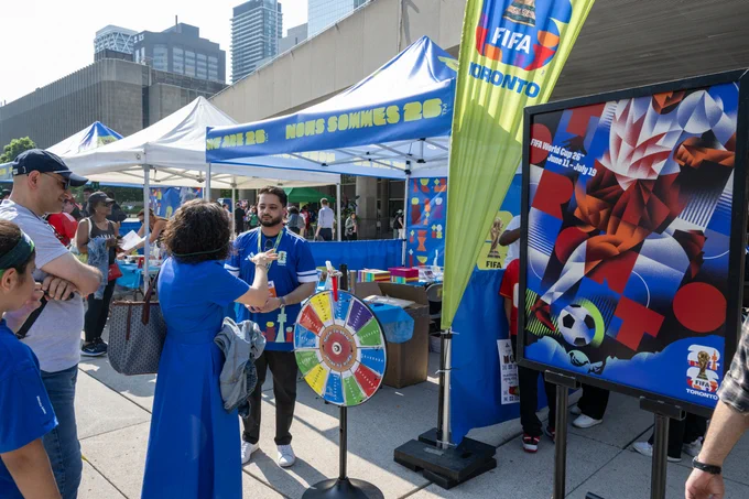 People gathered around a colorful prize wheel at an outdoor FIFA Toronto event with tents and city buildings in the background.