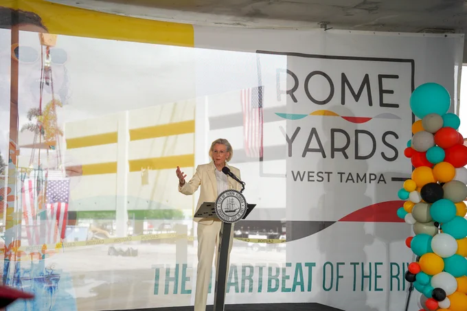 Mayor Jane Castor stands at a podium with a microphone, under a large canopy bearing the text "Rome Yards West Tampa." Colorful balloons decorate the side. The podium displays a seal that suggests an official event.