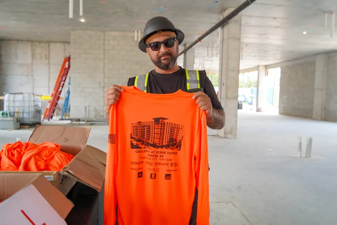 A person in a construction setting holds up an orange long-sleeve shirt. The shirt features a graphic of the Gallery at Rome Yards development. Nearby, there are boxes containing more shirts. The construction area has unfinished walls and visible piping on the ceiling.