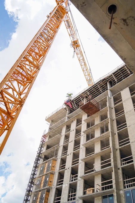 A tall construction crane positioned beside a multi-story building under construction. The building has unfinished concrete floors and several window openings. A partly cloudy sky is visible in the background.