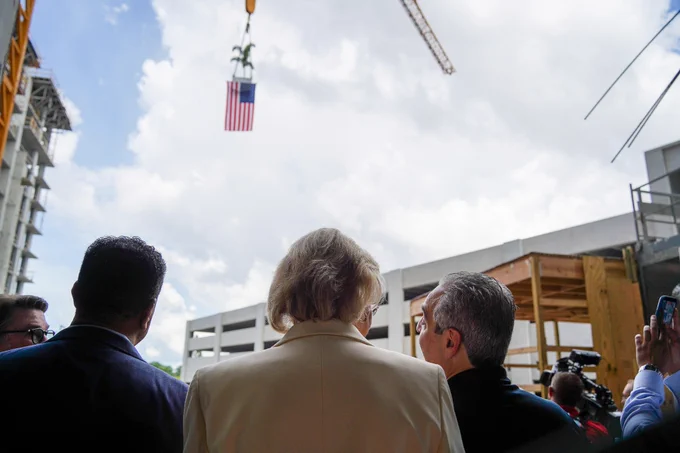 A group of people stand observing a construction site. A crane is hoisting a small tree and an American flag against a cloudy sky. Cameras and a few construction structures are visible.