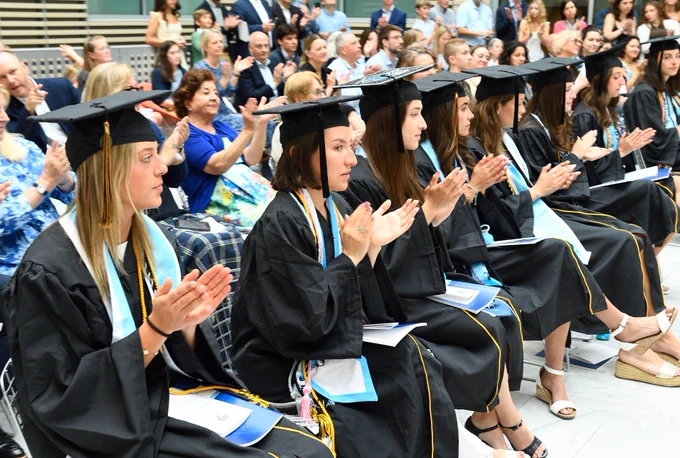 A group of graduates in caps and gowns sit in a row, applauding. The audience in the background is also seen clapping.