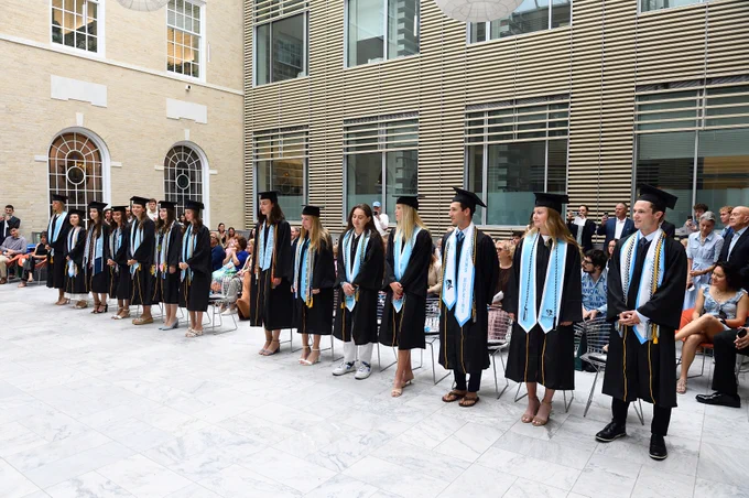 A group of graduates in caps and gowns stand in an open courtyard. They are lined up in front of a seated audience. The building features large windows and a beige brick exterior.