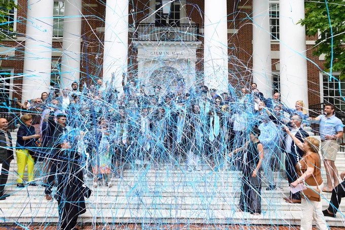 A large group of people celebrate on the steps of a building with white columns, surrounded by blue and white streamers in the air. The building has a sign that says "Gilman Hall."
