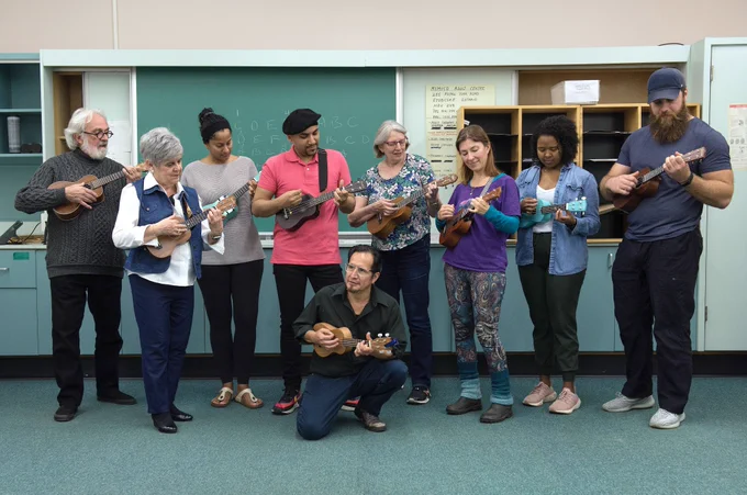 A group of adults standing in a classroom play ukuleles together in a line, with a chalkboard and shelving units behind them, while one person kneels in front also playing a ukulele.