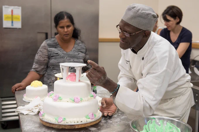 A chef carefully decorates a two-tier cake on a metal worktable, while others watch in a kitchen setting; baking tools and a bowl of green frosting are visible nearby.