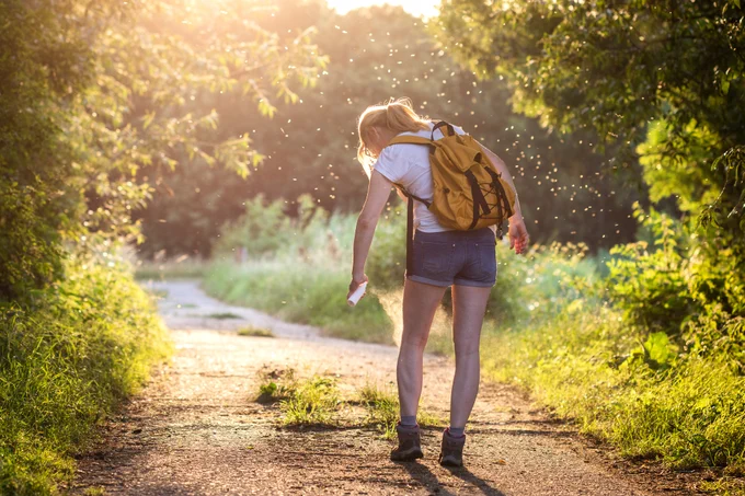 Young woman with a yellow backpack walking on a sunlit forest path spraying insect repellent