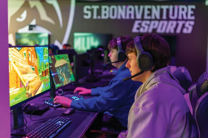 Two gamers wearing headsets intensely playing a colorful first-person shooter game at St. Bonaventure Esports gaming station.