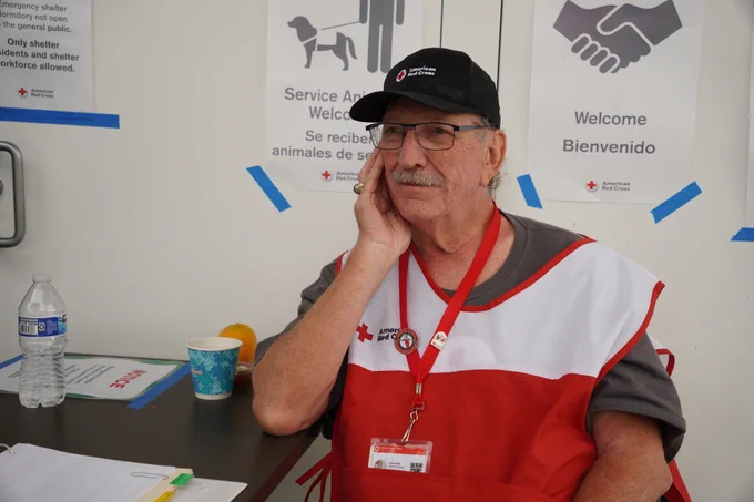 An individual wearing a Red Cross vest and cap is seated at a table with a thoughtful expression. There's a water bottle, fruit, and paper cup on the table. The backdrop features Red Cross posters with service animal and welcome information.
