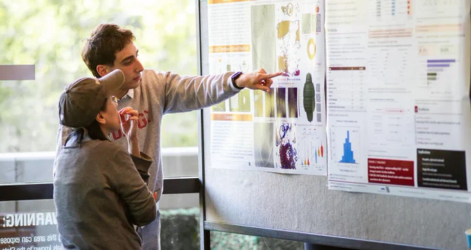 A researcher points to a poster describing his project while an onlooker listens. Photo by Ola Hopper.