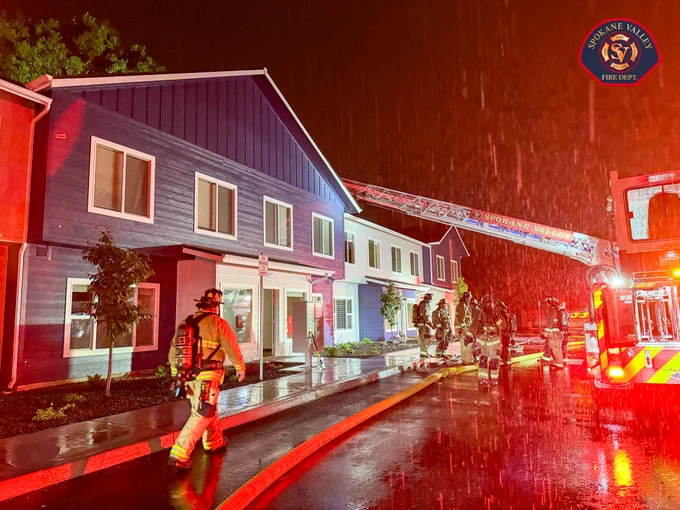 Firefighters and a fire truck from Spokane Valley Fire Department are responding to an emergency at a residential building at night. A ladder extends from the truck towards the building. Rain is visible in the scene.