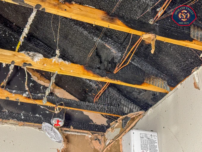 The image shows a damaged ceiling with exposed wooden beams and insulation, partially burnt and blackened. Tangled wires are visible, along with a damaged smoke detector hanging down. On the top right corner, a logo for the Spokane Valley Fire Department is present.