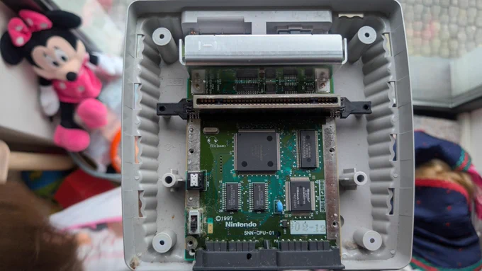 Super Famicom Jr. SHVC-101 opened up for the first time, original electrolytic capacitors and the ribbon cable connector visible, with dust accumulation on the board before cleaning.