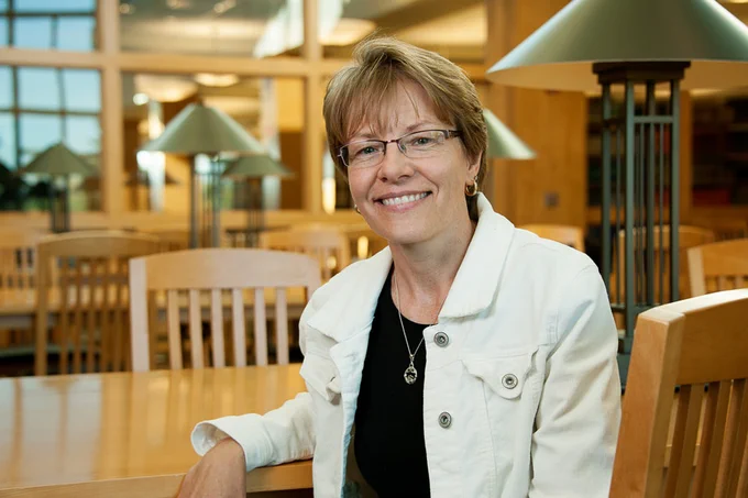 Woman sitting at a wooden library table smiling at the camera while wearing glasses, a white jacket, and black top.