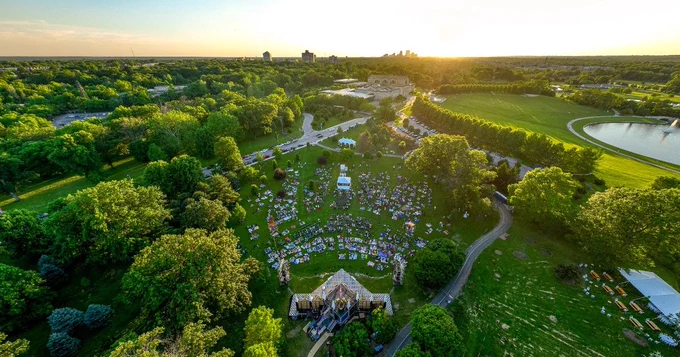 An aerial over Shakespeare Glen in Forest Park shows pathways and full-leaf tree lines create the edges of a diamond-like glen. The Saint Louis Art Museum is centered on top and a temporary stage sits at the bottom while a crowd of people sit on blankets throughout the green scape.
