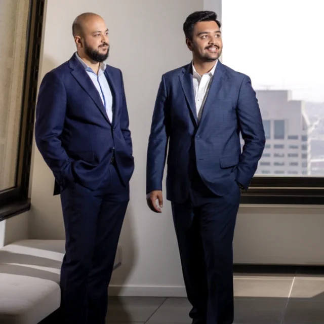 Two individuals in suits stand in an office space with a large window. One is seated on a bench, and the other stands nearby. A cityscape is visible through the window. The atmosphere appears professional.