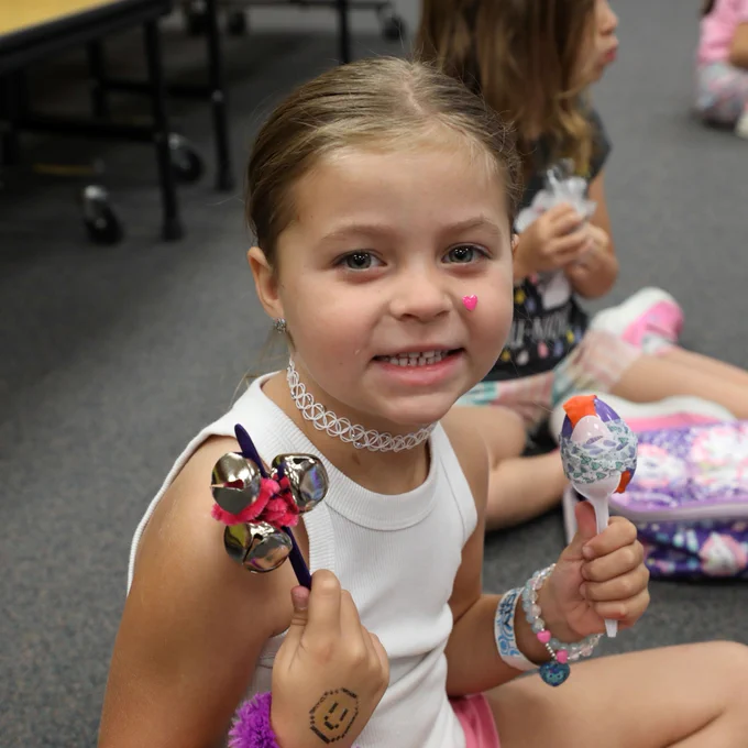 A child sitting on the floor in a casual setting holding two decorated musical instruments, one with bells and the other with colorful designs. Other children are in the background.