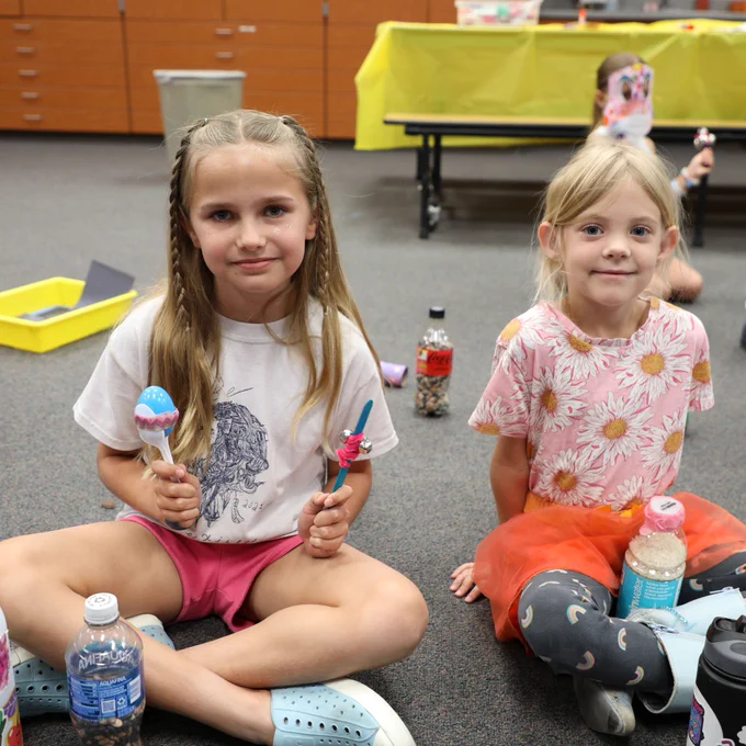 Two students are sitting on the floor of a classroom. One student is holding musical instruments she created.