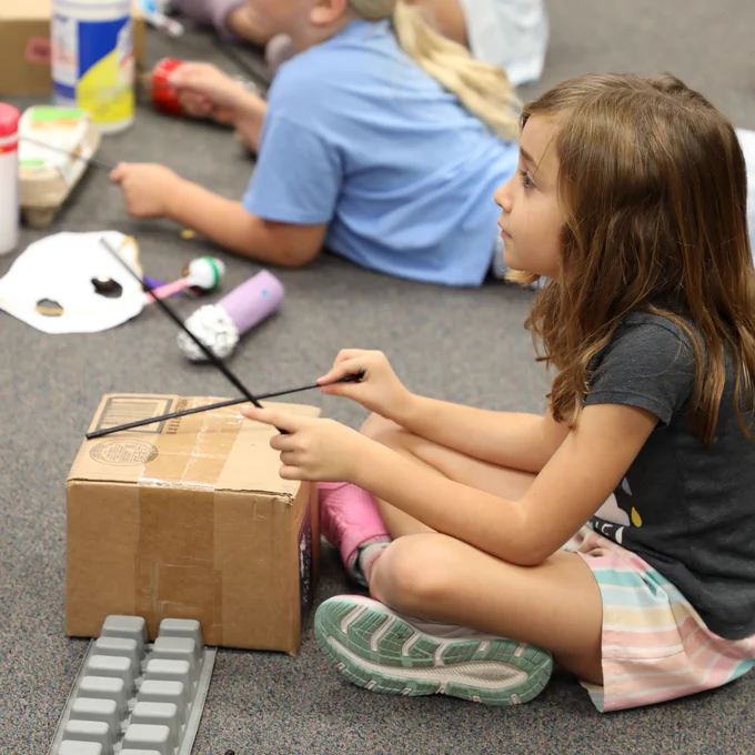 A young person in a classroom setting uses drumsticks to play on a cardboard box. Other children are in the background engaging in similar activities, with various musical instruments and materials scattered around.