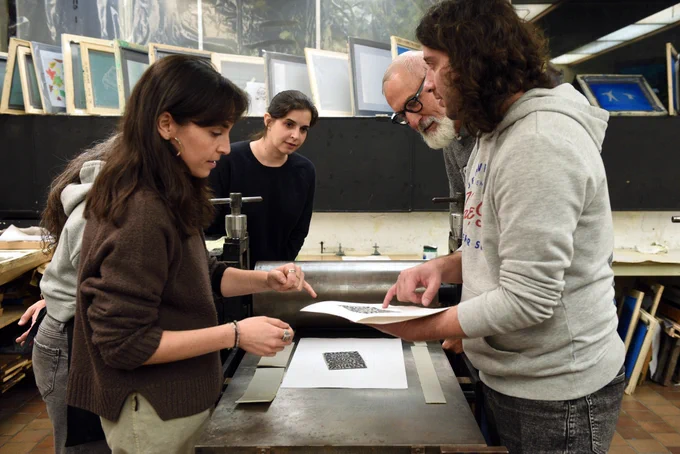 Several adults gather around a printing press in a workshop, closely examining and pointing at a printed sheet on the machine; framed prints and tools line the background shelves.