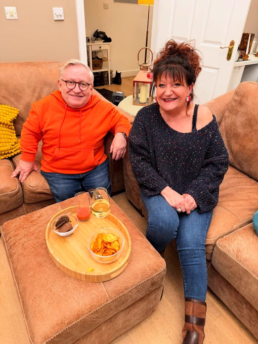 Simon in an orange hoodie and Jane in a navy jumper. Both sat on sofas looking up at the camera smiling. There’s the tray of nibbles in front of them.