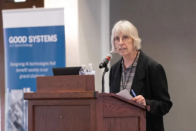 Sharon Strover speaks at a podium during the 2025 Good Systems Symposium. A Good Systems banner is visible in the background.