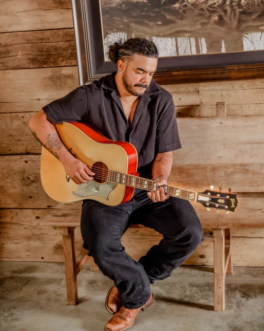 A person seated, playing an acoustic Gibson Custom Murphy Lab Dove guitar, in a room with wooden walls and a painting in the background. The individual is wearing a dark shirt and pants, with brown shoes.