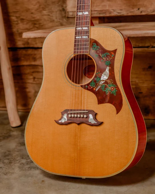 A Gibson acoustic Dove guitar with a light wood finish is resting upright. The guitar features an intricate pickguard design with flowers and a bird. The background includes a wooden bench and a rustic wooden wall.