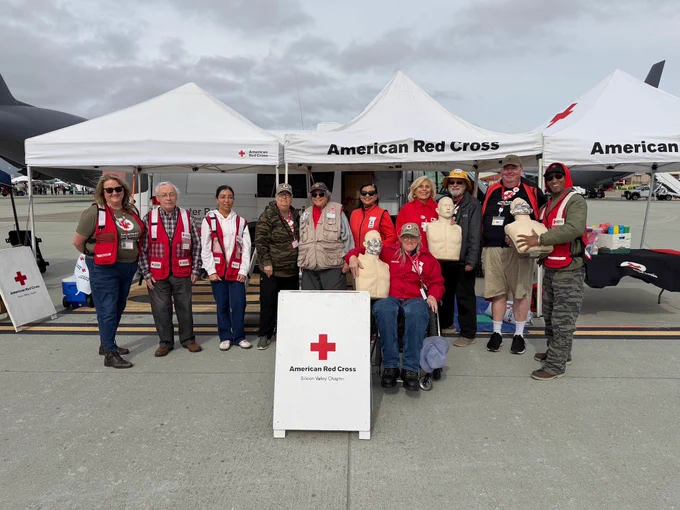 Group of people wearing American Red Cross vests stand in front of tents with American Red Cross signage. Some hold CPR mannequins, and they are positioned on an outdoor surface, such as a tarmac.