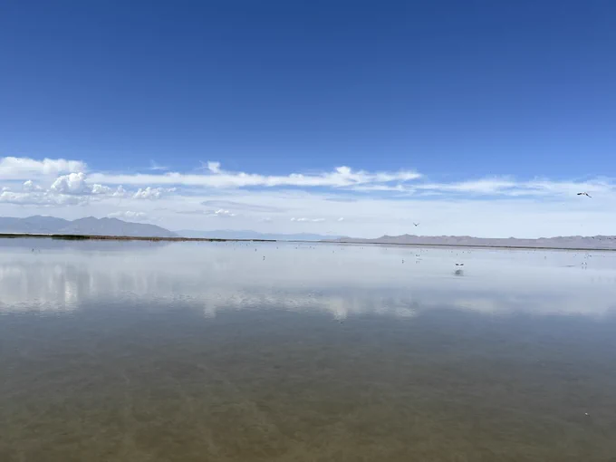 Calm lake reflecting a blue sky with scattered clouds and distant mountains on the horizon.