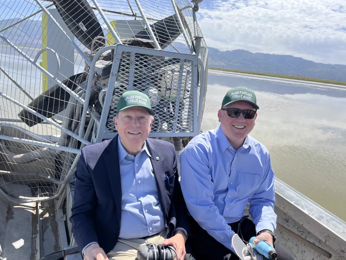 Two men sit on an airboat in a shallow lake or wetland under a bright blue sky with scattered clouds. Both are wearing green hats that read “MAKE THE LAKE GREAT AGAIN” and light blue button-down shirts. The man on the left wears a navy blazer and holds hearing protection, while the man on the right wears sunglasses and smiles toward the camera. A large airboat propeller and protective cage fill the background, with calm water and mountains visible in the distance.