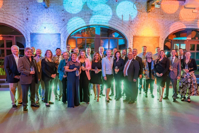 A group of people, including UTEP President Heather Wilson and honorees at the 2026 Miner Business Impact Awards, dressed in formal attire stand smiling inside an event space with exposed brick walls, large arched windows, and colorful lights.