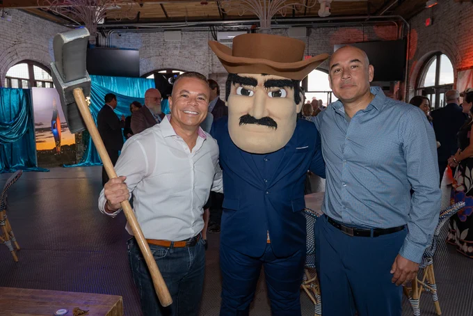 A person holding an axe, a person in a costume with a large hat and mustache, and another person standing together indoors at an event. The background includes brick walls, draped fabric, and chairs.