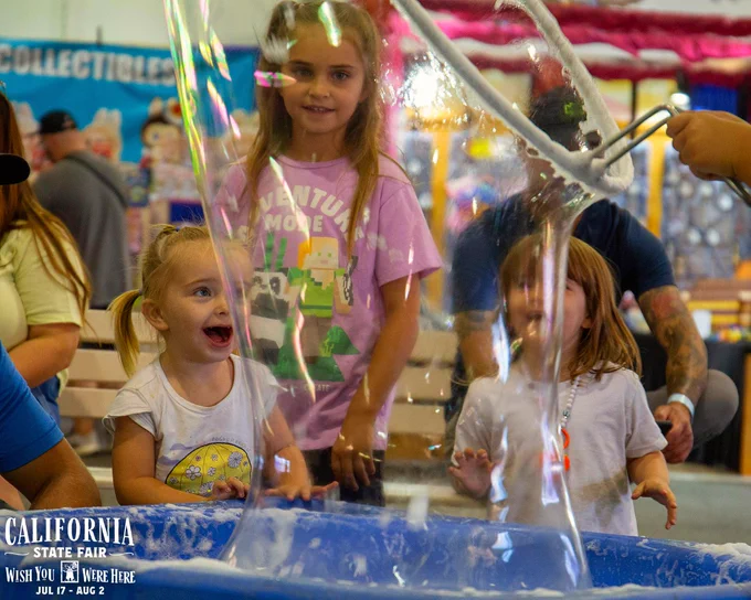Children at the California State Fair captivated by a giant bubble demonstration inside a booth filled with colorful fair decorations.