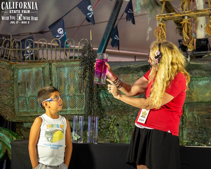 A woman in a red shirt shows a tall glass with pink liquid to a young boy at the California State Fair.
